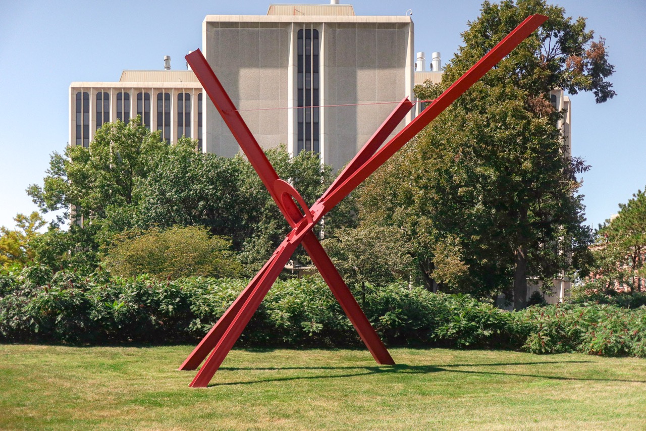 Red metal abstract sculpture on green lawn, with tall buildings and trees in the background.
