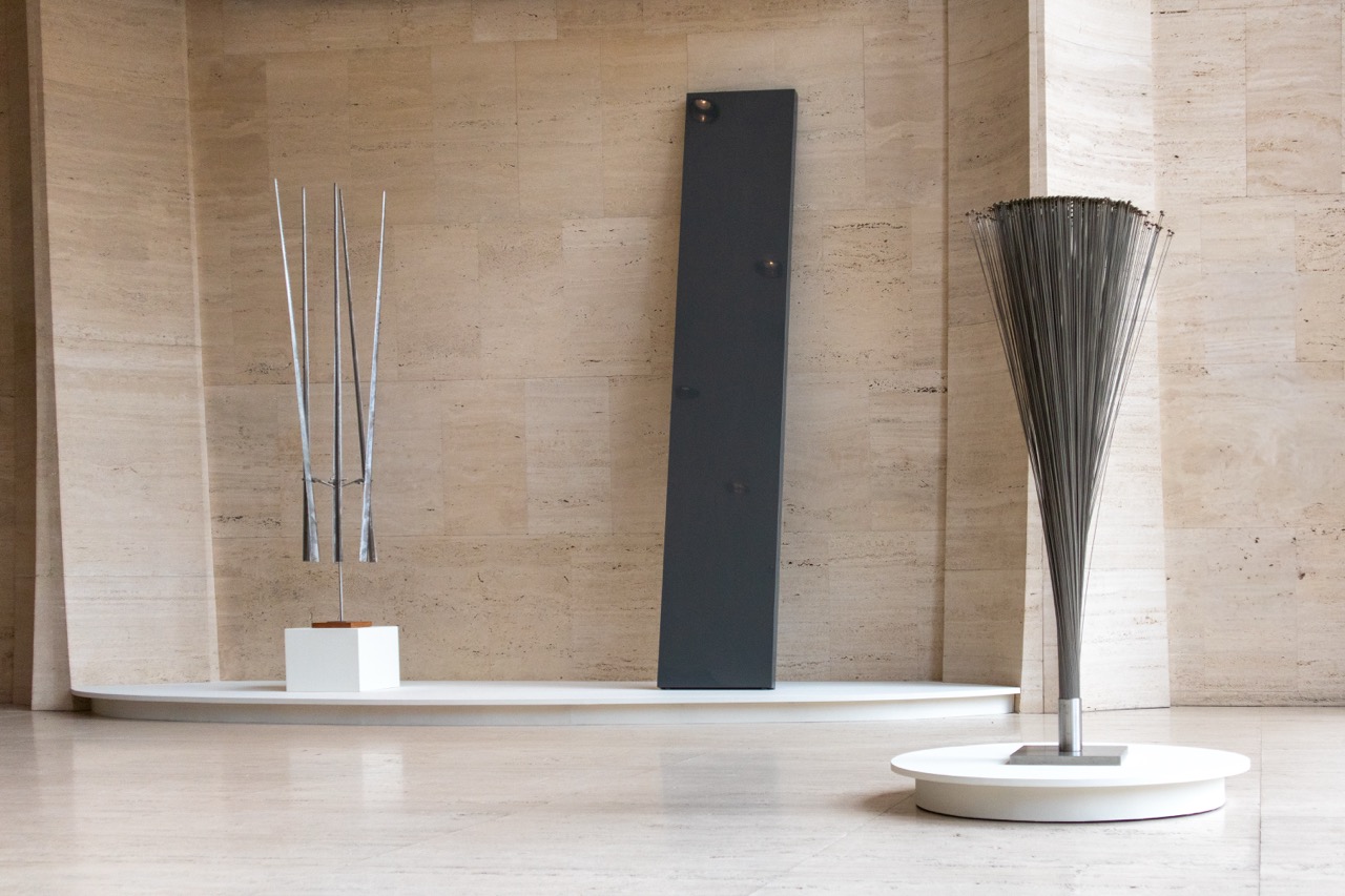 Three minimalist sculptures installed on white platforms in a travertine room.