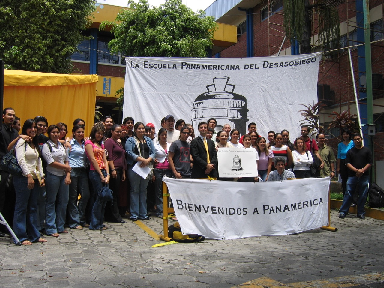 A photo of a large people smiling in front of a banner that reads "La Escuela Panamericana Del Desasosiego"